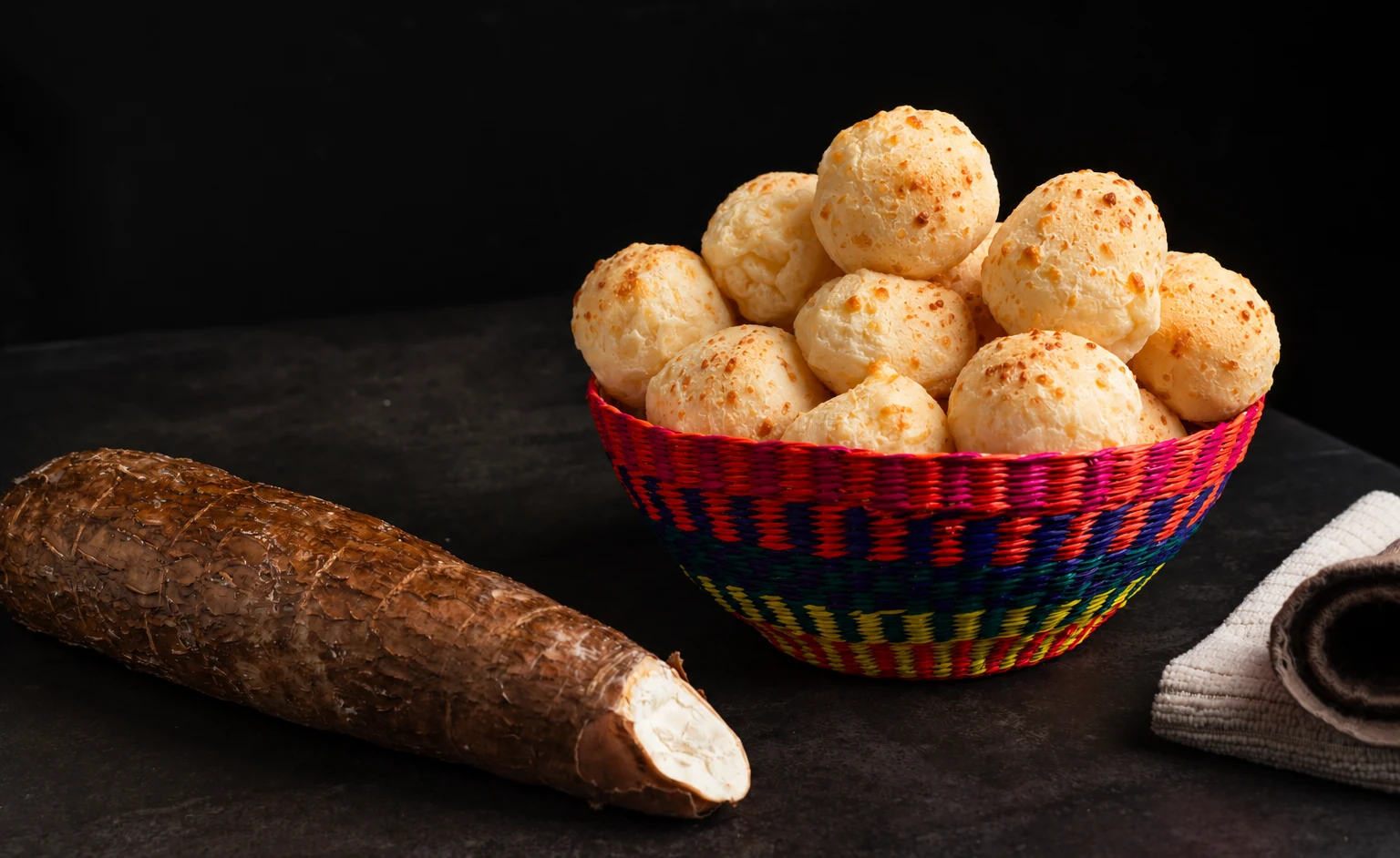 Cesta de colores llena de pan de yuca con queso, junto a una raíz de yuca